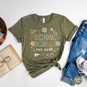 Peut inclure: T-shirt vert olive avec l'inscription "SCHOOL SECRETARY mrs. smith" dans une police de caract&egrave;res color&eacute;e et ludique. Le motif comprend un avion en papier, un crayon, des fleurs et une pomme. Le t-shirt est associ&eacute; &agrave; un jean bleu et des baskets blanches.