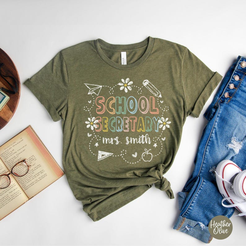Peut inclure: T-shirt vert olive avec l'inscription "SCHOOL SECRETARY mrs. smith" dans une police de caract&egrave;res color&eacute;e et ludique. Le motif comprend un avion en papier, un crayon, des fleurs et une pomme. Le t-shirt est associ&eacute; &agrave; un jean bleu et des baskets blanches.