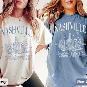 K&ouml;nnte beinhalten: Zwei T-Shirts mit einem Nashville-Design. Das Design zeigt eine Flasche Alkohol, eine Gitarre, einen Cowboyhut und Stiefel. Der Text auf den Shirts lautet "Nashville Social & Cocktail Club Alyssa's Bachelorette" mit dem Jahr 2024. Das Shirt auf der linken Seite ist elfenbeinfarben und das Shirt auf der rechten Seite ist jeansblau.