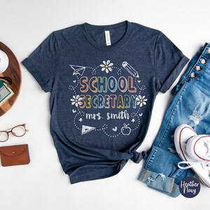 Peut inclure: T-shirt bleu marine avec l'inscription "SCHOOL SECRETARY mrs. smith" en lettres et graphismes color&eacute;s. Le motif comprend un avion en papier, des fleurs, un crayon, une r&egrave;gle, une pomme et des &eacute;toiles.
