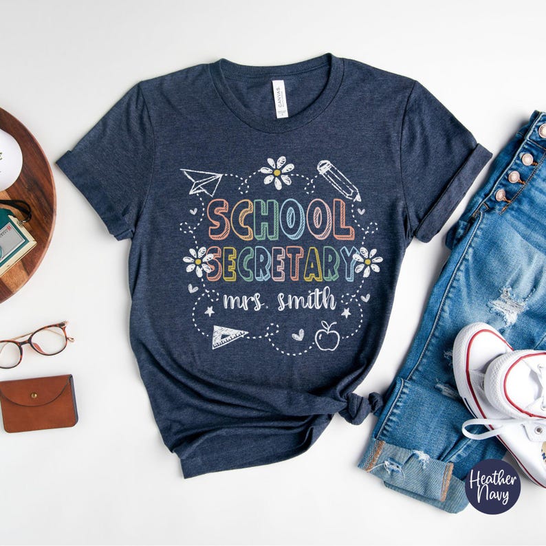 Peut inclure: T-shirt bleu marine avec l'inscription "SCHOOL SECRETARY mrs. smith" en lettres et graphismes color&eacute;s. Le motif comprend un avion en papier, des fleurs, un crayon, une r&egrave;gle, une pomme et des &eacute;toiles.
