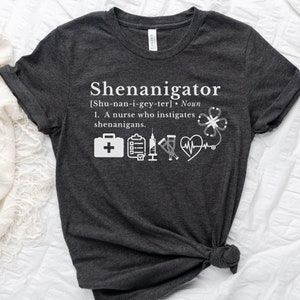 May include: Dark gray t-shirt with white text that reads "Shenanigator [Shu-nan-i-gey-ter] Noun 1. A nurse who instigates shenanigans."  The shirt also features a white medical cross, clipboard, syringe, stethoscope, and heart with a heartbeat line.