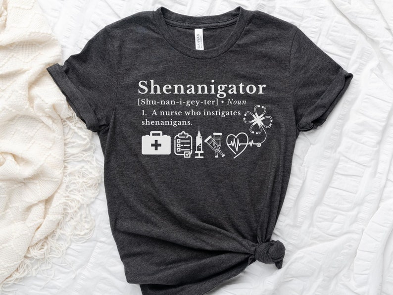 May include: Dark gray t-shirt with white text that reads "Shenanigator [Shu-nan-i-gey-ter] Noun 1. A nurse who instigates shenanigans."  The shirt also features a white medical cross, clipboard, syringe, stethoscope, and heart with a heartbeat line.