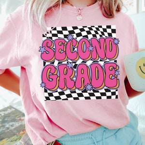 以下が含まれることがあります： 黒と白の市松模様の背景に、ピンクと紫の文字で「SECOND GRADE」と書かれたピンクのTシャツです。