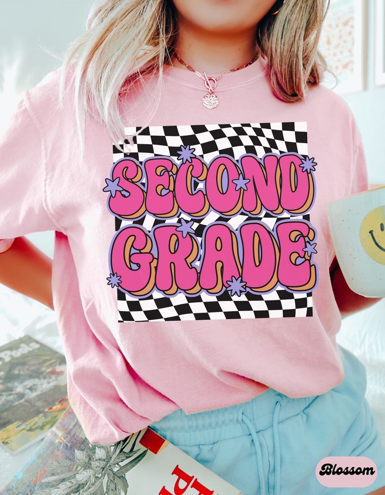以下が含まれることがあります： 黒と白の市松模様の背景に、ピンクと紫の文字で「SECOND GRADE」と書かれたピンクのTシャツです。