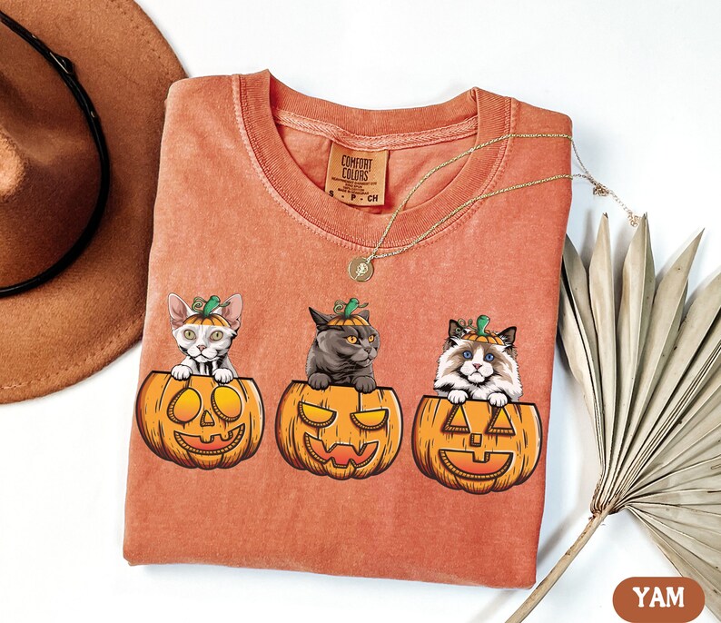 May include: A burnt orange t-shirt featuring three cats wearing jack-o-lantern hats. The cats are sitting inside the jack-o-lanterns. The text "COMFORT COLORS" is on the shirt.