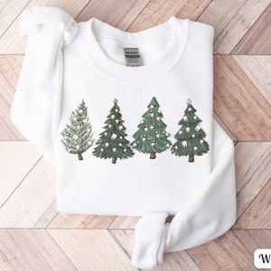 K&ouml;nnte beinhalten: Wei&szlig;er Pullover mit einem Motiv von f&uuml;nf Weihnachtsb&auml;umen, die mit Z&auml;hnen verziert sind. Die B&auml;ume sind gr&uuml;n mit wei&szlig;en Z&auml;hnen und goldenen Sternen.