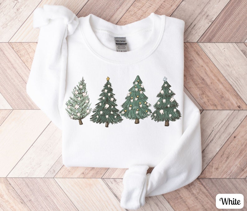 K&ouml;nnte beinhalten: Wei&szlig;er Pullover mit einem Motiv von f&uuml;nf Weihnachtsb&auml;umen, die mit Z&auml;hnen verziert sind. Die B&auml;ume sind gr&uuml;n mit wei&szlig;en Z&auml;hnen und goldenen Sternen.