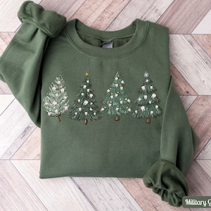 K&ouml;nnte beinhalten: Ein gr&uuml;nes Sweatshirt mit einem Weihnachtsbaum-Design, das Z&auml;hne als Ornamente zeigt. Die B&auml;ume sind mit goldenen Sternen und Schneeflocken verziert. Das Sweatshirt ist in Milit&auml;rgr&uuml;n gehalten.