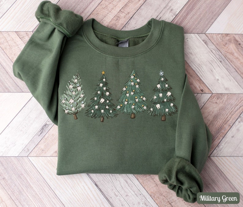 K&ouml;nnte beinhalten: Ein gr&uuml;nes Sweatshirt mit einem Weihnachtsbaum-Design, das Z&auml;hne als Ornamente zeigt. Die B&auml;ume sind mit goldenen Sternen und Schneeflocken verziert. Das Sweatshirt ist in Milit&auml;rgr&uuml;n gehalten.