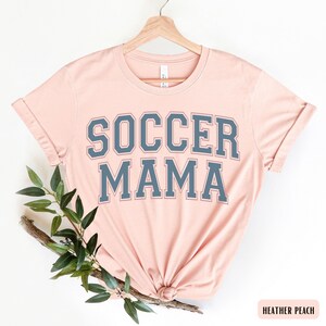 May include: A heather peach t-shirt with the text "SOCCER MAMA" in a navy blue varsity-style font.