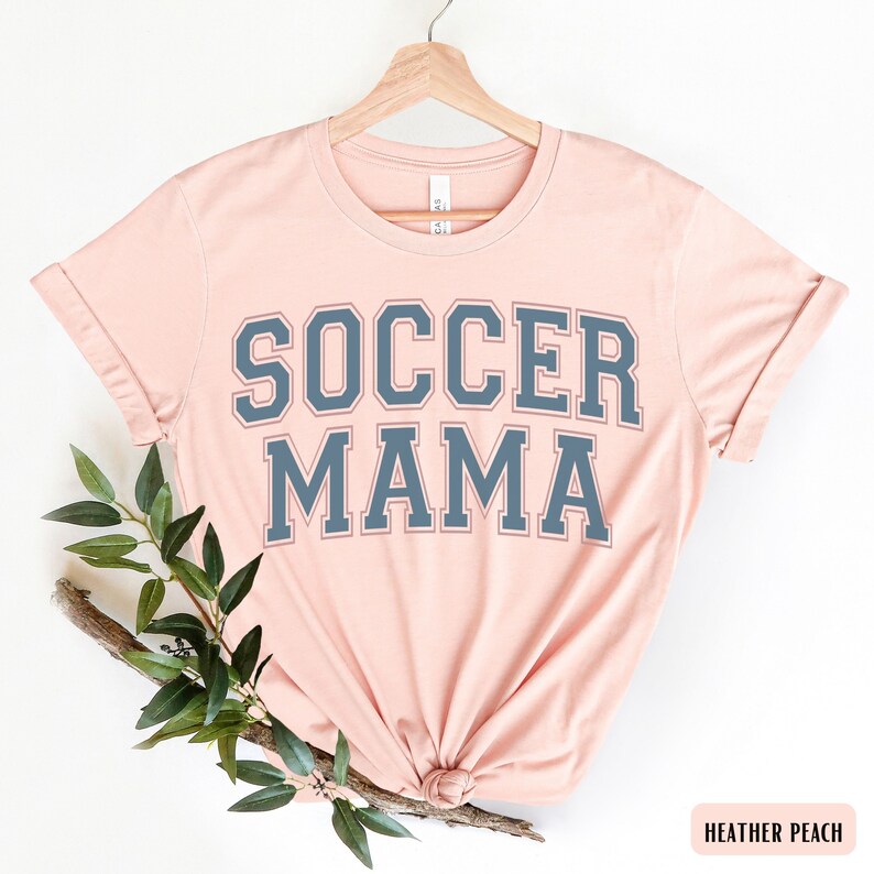 May include: A heather peach t-shirt with the text "SOCCER MAMA" in a navy blue varsity-style font.