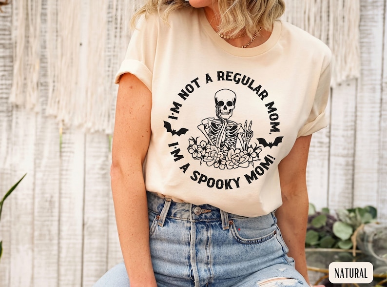 May include: A beige t-shirt with a black graphic of a skeleton holding a peace sign surrounded by flowers and bats. The text on the shirt reads "I'm not a regular mom. I'm a spooky mom!"
