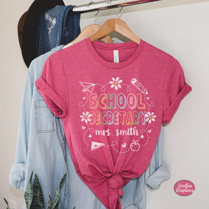 Peut inclure: Un t-shirt rose avec l'inscription "SCHOOL SECRETARY mrs. smith" en lettres color&eacute;es et des &eacute;l&eacute;ments d&eacute;coratifs. Le motif comprend un avion en papier, un crayon, des fleurs et une pomme. Le t-shirt est suspendu &agrave; un cintre en bois.