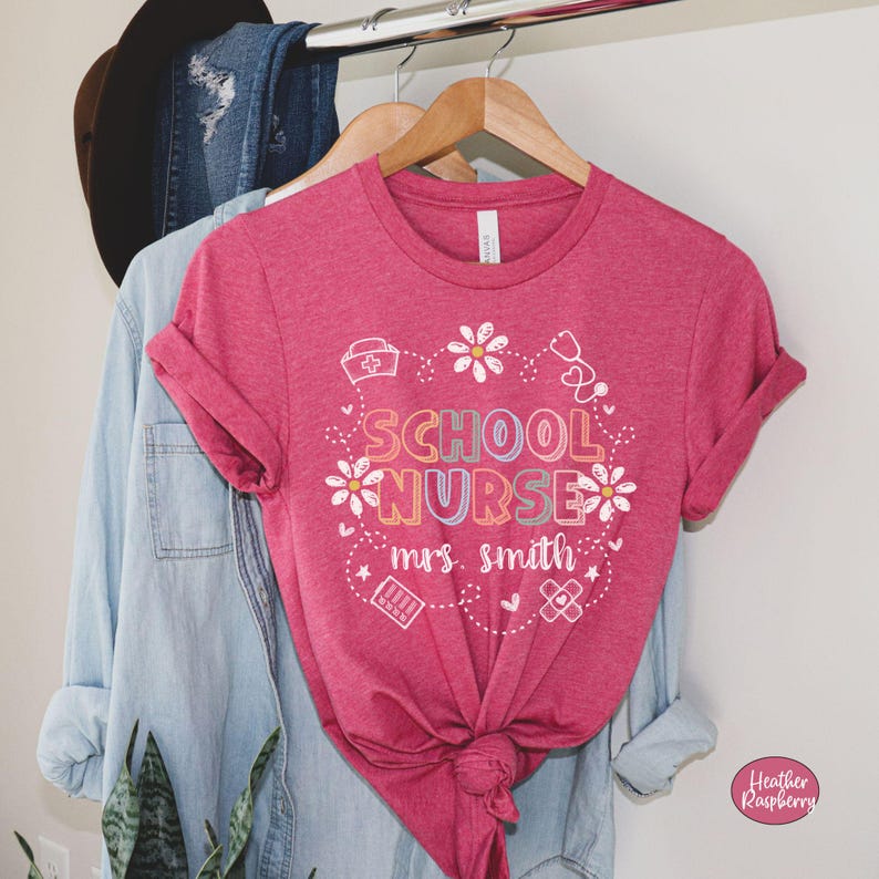 Puede incluir: Una camiseta de color frambuesa con las palabras "SCHOOL NURSE" en letras coloridas, rodeadas de margaritas blancas y s&iacute;mbolos m&eacute;dicos. El nombre "mrs. smith" est&aacute; impreso debajo. La camiseta est&aacute; colgada en una percha de madera.