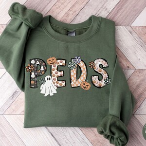 May include: A military green sweatshirt with the word "PEDS" in a decorative font. The letters are filled with Halloween-themed patterns, including ghosts, pumpkins, and flowers. The sweatshirt has long sleeves and a crew neck.