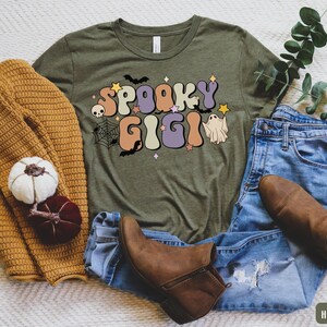 May include: A green t-shirt with the text "Spooky Gigi" in a retro font. The text is decorated with bats, a skull, a spiderweb, and a ghost. The shirt is laid out on a white surface with a brown sweater, blue jeans, and brown boots.