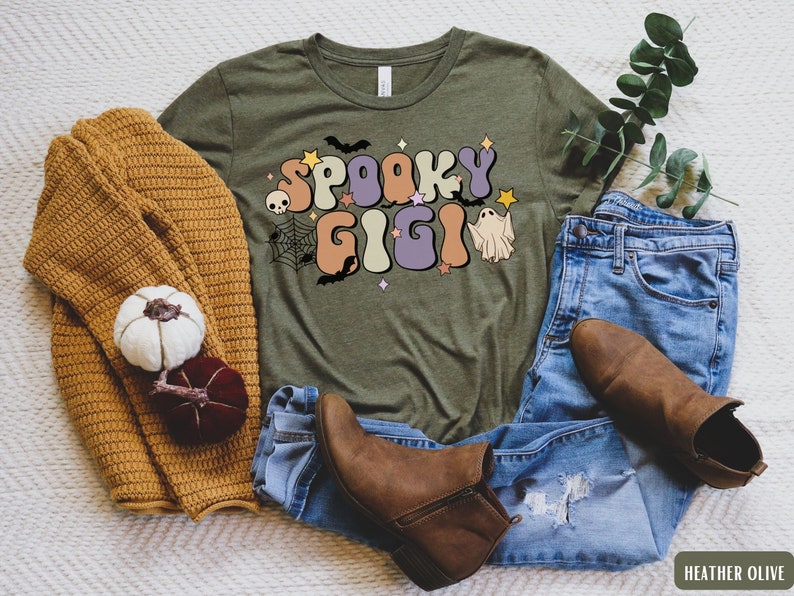 May include: A green t-shirt with the text "Spooky Gigi" in a retro font. The text is decorated with bats, a skull, a spiderweb, and a ghost. The shirt is laid out on a white surface with a brown sweater, blue jeans, and brown boots.