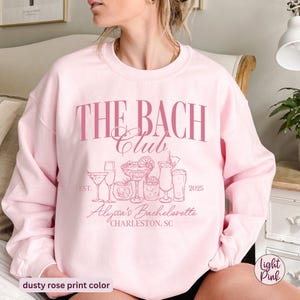 K&ouml;nnte beinhalten: Rosafarbener Pullover mit dem Text "THE BACH Club" und einer Grafik von Cocktails. Der Text "EST. 2025" und "Alyssa's Bachelorette CHARLESTON, SC" ist ebenfalls auf dem Pullover aufgedruckt.