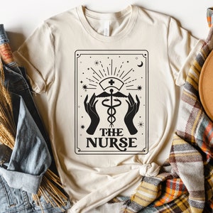 May include: A beige t-shirt featuring a black and white graphic design of a tarot card. The card depicts a medical caduceus symbol with the text "The Nurse" underneath.