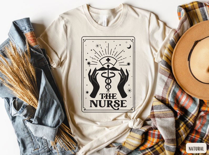 May include: A beige t-shirt featuring a black and white graphic design of a tarot card. The card depicts a medical caduceus symbol with the text "The Nurse" underneath.