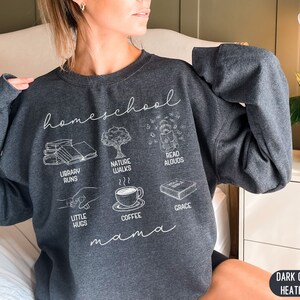 Puede incluir: Sudadera de color gris oscuro jaspeado con el texto "homeschool mama" e ilustraciones de libros, paseos por la naturaleza, lectura en voz alta, peque&ntilde;os abrazos, caf&eacute; y gracia.
