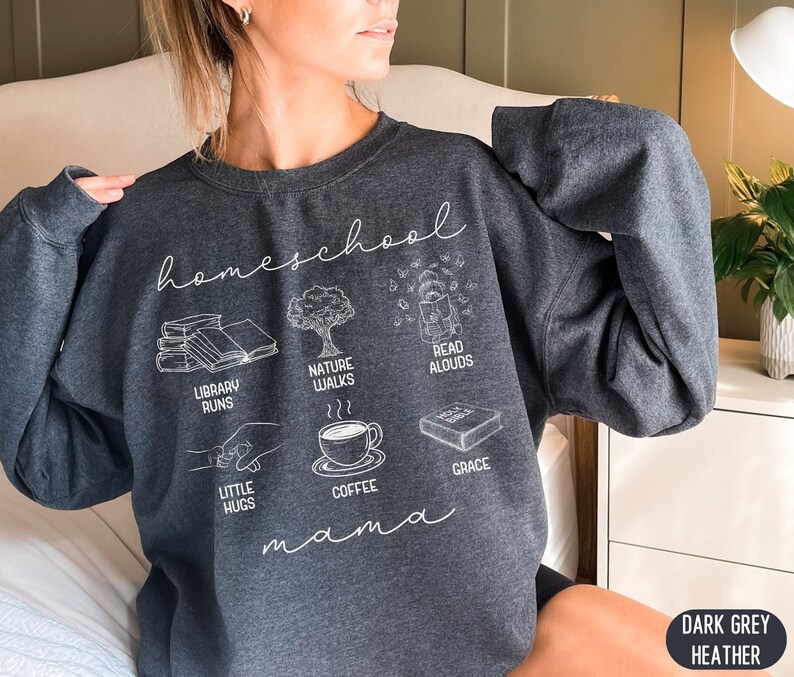 Puede incluir: Sudadera de color gris oscuro jaspeado con el texto "homeschool mama" e ilustraciones de libros, paseos por la naturaleza, lectura en voz alta, peque&ntilde;os abrazos, caf&eacute; y gracia.