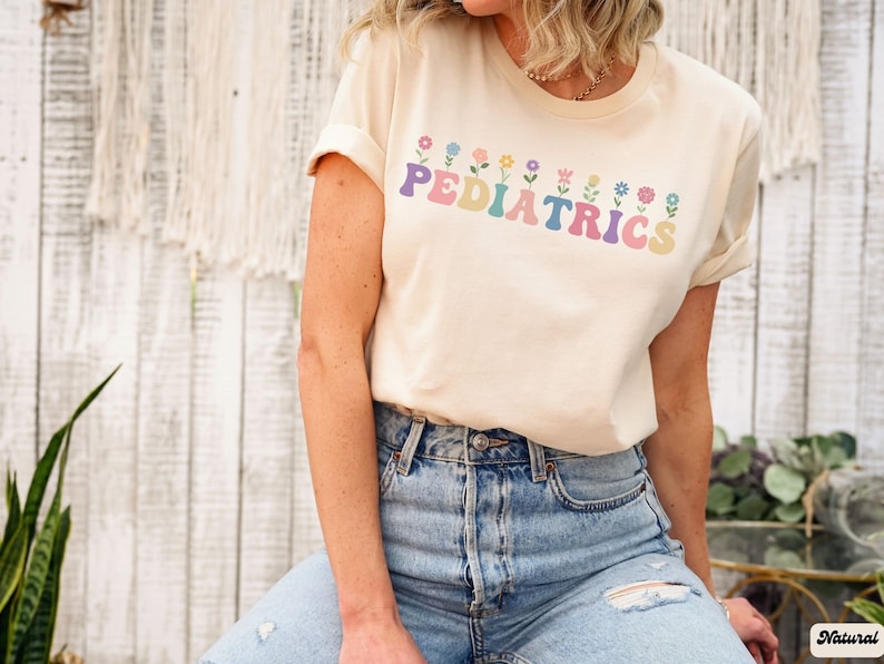 Puede incluir: Una mujer lleva una camiseta beige claro con el texto "PEDIATRICS" en letras de colores con flores encima y debajo del texto. Lleva unos vaqueros azules.