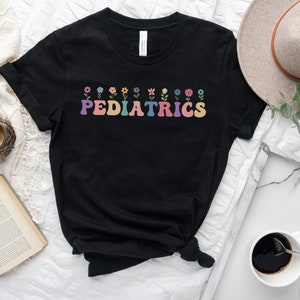 Puede incluir: Camiseta negra con un gr&aacute;fico de "PEDIATRICS" de colores y una fila de flores sobre el texto.