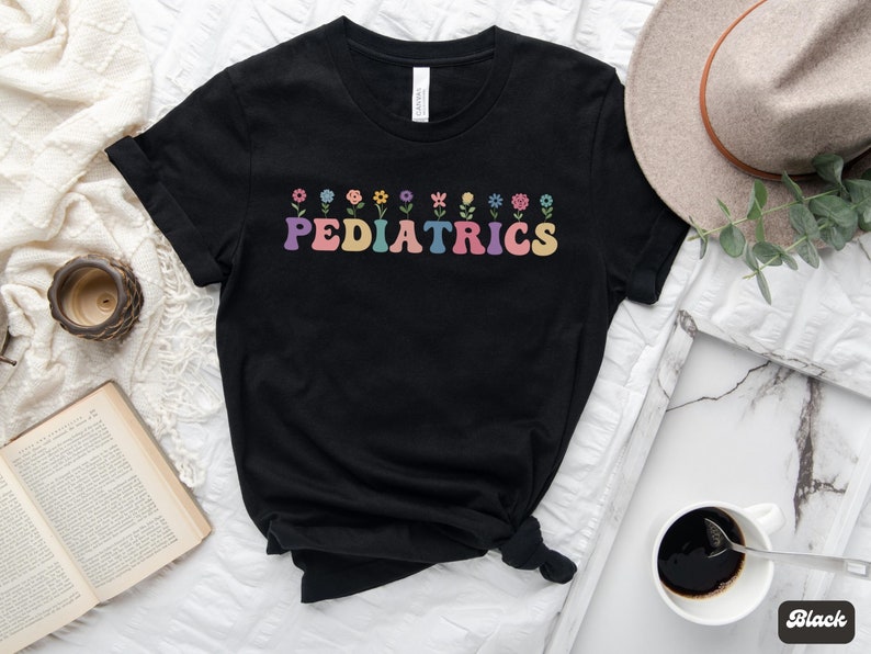 Puede incluir: Camiseta negra con un gr&aacute;fico de "PEDIATRICS" de colores y una fila de flores sobre el texto.