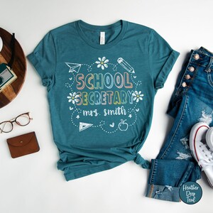 Peut inclure: T-shirt de couleur teal avec l'inscription "SCHOOL SECRETARY mrs. smith" dans une police fantaisiste et color&eacute;e. Le motif comprend un avion en papier, un crayon, des fleurs et une pomme. Le t-shirt est associ&eacute; &agrave; un jean bleu et des baskets blanches.