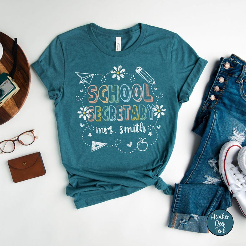 Peut inclure: T-shirt de couleur teal avec l'inscription "SCHOOL SECRETARY mrs. smith" dans une police fantaisiste et color&eacute;e. Le motif comprend un avion en papier, un crayon, des fleurs et une pomme. Le t-shirt est associ&eacute; &agrave; un jean bleu et des baskets blanches.