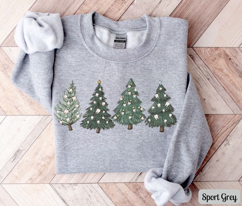 K&ouml;nnte beinhalten: Ein graues Sweatshirt mit einem Weihnachtsbaum-Design, das Z&auml;hne als Schmuck verwendet. Die B&auml;ume sind gr&uuml;n mit wei&szlig;en Z&auml;hnen und gelben Sternen. Das Sweatshirt ist in Sportgrau gehalten.