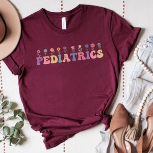 Puede incluir: Una camiseta color burdeos con un dise&ntilde;o floral y el texto "PEDIATRICS" en letras de colores.