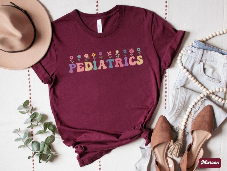 Puede incluir: Una camiseta color burdeos con un dise&ntilde;o floral y el texto "PEDIATRICS" en letras de colores.