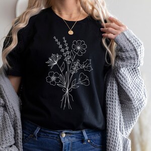 以下が含まれることがあります： 白い線で描かれた花束がプリントされた黒いTシャツ。花束には、さまざまな花、葉、茎が含まれています。