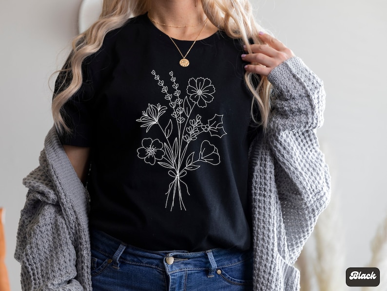 以下が含まれることがあります： 白い線で描かれた花束がプリントされた黒いTシャツ。花束には、さまざまな花、葉、茎が含まれています。
