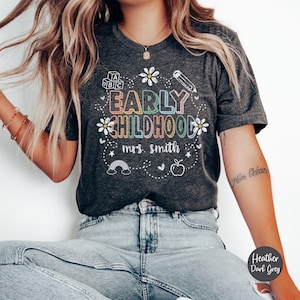Pode incluir: T-shirt cinza escuro mesclado com as palavras "EARLY CHILDHOOD mrs. smith" em um design colorido e caprichoso. O design inclui um arco-&iacute;ris, uma ma&ccedil;&atilde;, um l&aacute;pis e flores. A camisa &eacute; cinza escuro.