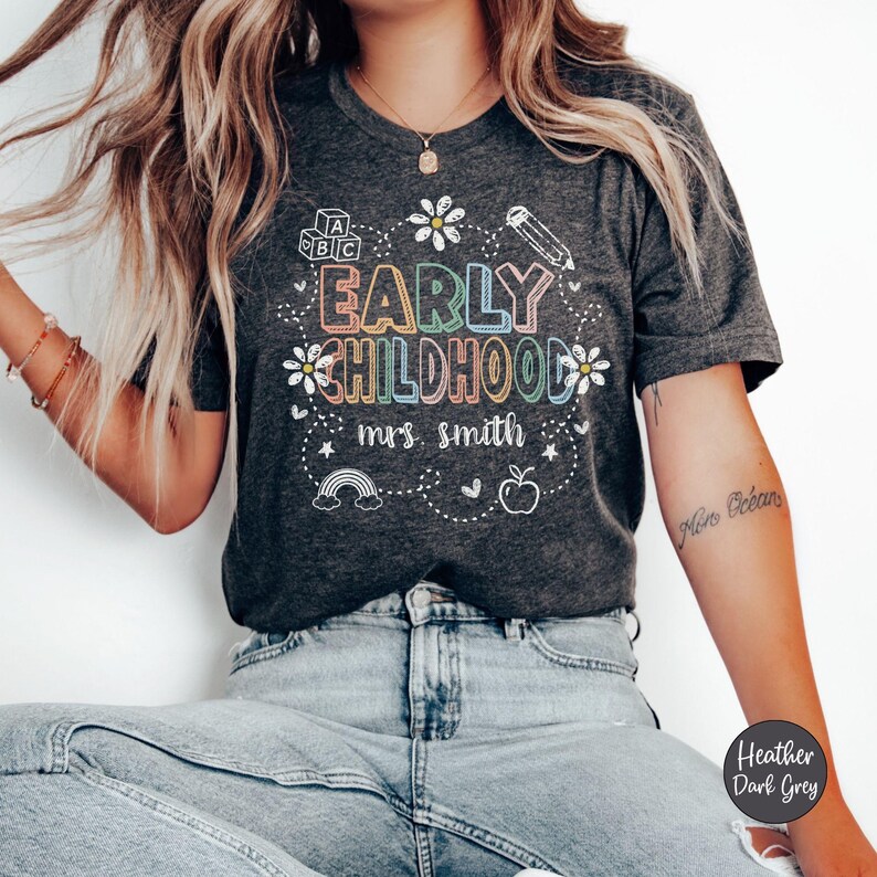 Pode incluir: T-shirt cinza escuro mesclado com as palavras "EARLY CHILDHOOD mrs. smith" em um design colorido e caprichoso. O design inclui um arco-&iacute;ris, uma ma&ccedil;&atilde;, um l&aacute;pis e flores. A camisa &eacute; cinza escuro.