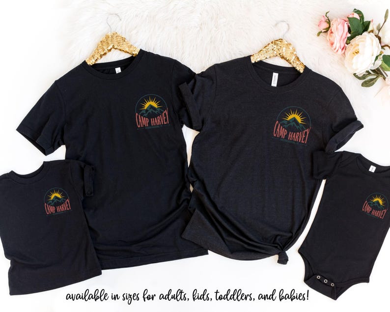 K&ouml;nnte beinhalten: Zwei schwarze T-Shirts mit kurzen &Auml;rmeln, ein Kinder-T-Shirt und ein Baby-Body. Jedes Kleidungsst&uuml;ck zeigt auf der Brust ein kreisf&ouml;rmiges Motiv mit einer gelben Sonne &uuml;ber dunklen Bergen und dem darunter gedruckten Text Camp Harvey.