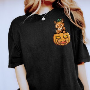 May include: Black t-shirt with a graphic of a cat inside a jack-o'-lantern. The cat is orange and white with green leaves on its head. The t-shirt has a crew neck and short sleeves.