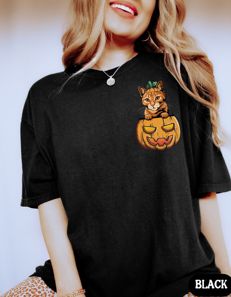 May include: Black t-shirt with a graphic of a cat inside a jack-o'-lantern. The cat is orange and white with green leaves on its head. The t-shirt has a crew neck and short sleeves.
