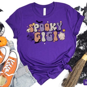 May include: Purple t-shirt featuring a graphic of the text "Spooky Gigi" in a retro font with a ghost and bats.
