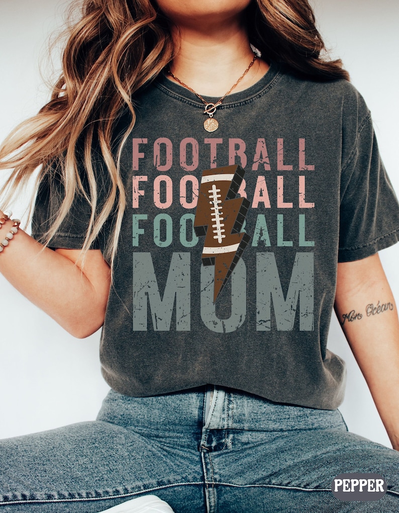 Op de afbeelding: Een grijs T-shirt met de tekst "FOOTBALL FOOTBALL FOOTBALL MOM" in roze, groen en grijs met een bliksem en een voetbalafbeelding.