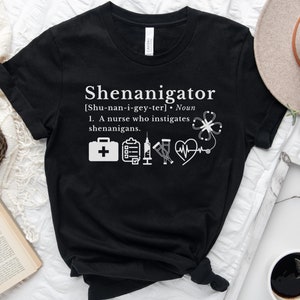 May include: Black t-shirt with white text that reads "Shenanigator [Shu-nan-i-gey-ter]. Noun 1. A nurse who instigates shenanigans."  The shirt also features a white medical symbol with a stethoscope, a heart rate monitor, a syringe, a clipboard, a first aid kit, and a four-leaf clover.