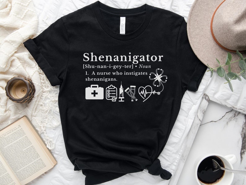 May include: Black t-shirt with white text that reads "Shenanigator [Shu-nan-i-gey-ter]. Noun 1. A nurse who instigates shenanigans."  The shirt also features a white medical symbol with a stethoscope, a heart rate monitor, a syringe, a clipboard, a first aid kit, and a four-leaf clover.