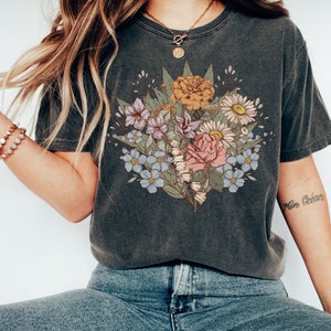 Pode incluir: T-shirt cinzento escuro com um design de buqu&ecirc; floral em tons de rosa, amarelo, azul e roxo. O buqu&ecirc; inclui rosas e margaridas. A camiseta &eacute; combinada com jeans azuis. A tatuagem no bra&ccedil;o diz "Mon Oc&eacute;an".