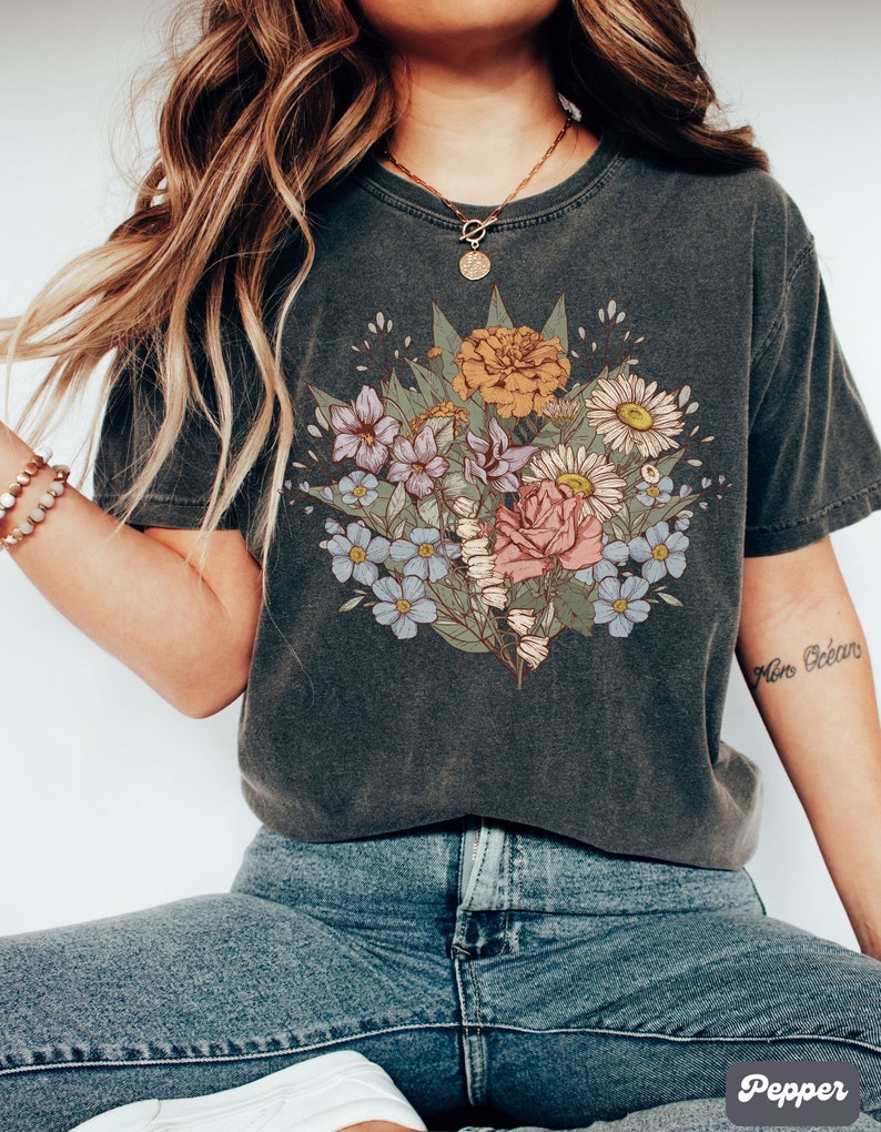Pode incluir: T-shirt cinzento escuro com um design de buqu&ecirc; floral em tons de rosa, amarelo, azul e roxo. O buqu&ecirc; inclui rosas e margaridas. A camiseta &eacute; combinada com jeans azuis. A tatuagem no bra&ccedil;o diz "Mon Oc&eacute;an".