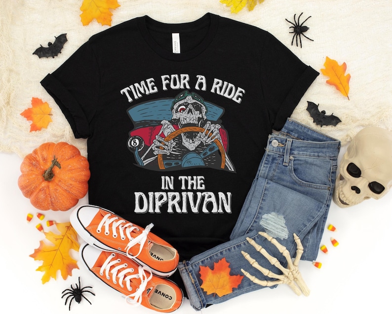May include: Black t-shirt with a skeleton driving a car and the text "Time for a ride in the dip-rivan". The shirt is paired with blue jeans, a skeleton hand, and orange and brown fall leaves.
