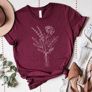 K&ouml;nnte beinhalten: Ein maroonfarbenes T-Shirt mit einer wei&szlig;en Linienzeichnung eines Blumenstrau&szlig;es. Die Blumen umfassen eine gro&szlig;e Pfingstrose und kleinere Lavendelzweige.